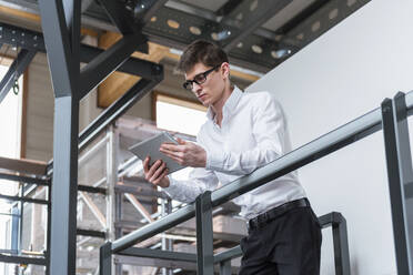 Selbstbewusster Geschäftsmann, der ein digitales Tablet benutzt, während er an einem Geländer in einer Fabrik steht - DIGF12702