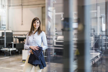 Lächelnde weibliche Fachkraft, die einen Blazer hält, während sie in einer Fabrik steht - DIGF12739