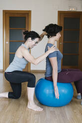Weibliche Physiotherapeutin hilft einem Patienten, der auf einem Fitnessball im Fitnessstudio sitzt - XLGF00212