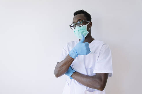 Afro-Arzt mit chirurgischer Maske zeigt Daumen hoch, während er vor weißem Hintergrund steht - JCMF00857