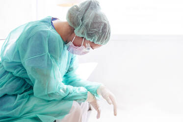 Sad female doctor wearing protective workwear sitting against window in hospital - JCMF00878