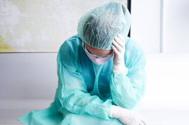 Sad female doctor wearing protective workwear sitting against wall in hospital - JCMF00881