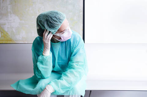 Sad doctor wearing protective workwear sitting against wall in hospital stock photo