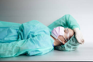 Tired female doctor wearing protective workwear resting on bench in hospital - JCMF00884