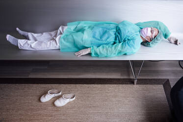 Tired female doctor wearing protective workwear lying on bench in hospital - JCMF00885