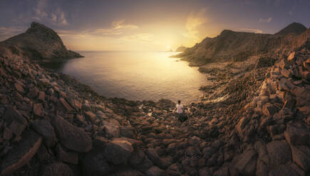 Spanien, Provinz Almeria, Frau sitzt allein am felsigen Küstenstrand im Naturpark Cabo de Gata-Nijar bei Sonnenuntergang - DVGF00092