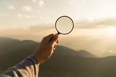 Close-Up Of Hand Holding Magnifying Glass Over Landscape Against Sky During Sunset - EYF06865