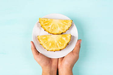 Cropped Image Of Hand Holding Pineapple Slices In Plate Over Blue Background - EYF06886