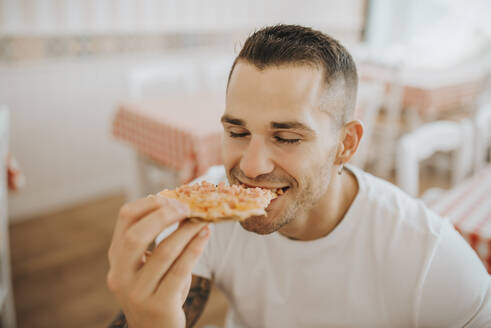 Nahaufnahme eines jungen Mannes mit geschlossenen Augen, der in einem Restaurant sitzt und Pizza isst - MIMFF00057