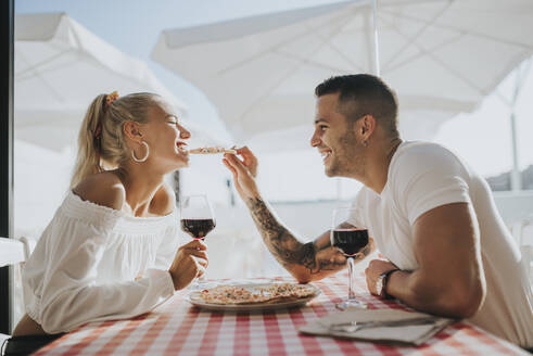 Glücklicher junger Mann füttert seine Freundin mit Pizza, während er im Restaurant sitzt - MIMFF00058
