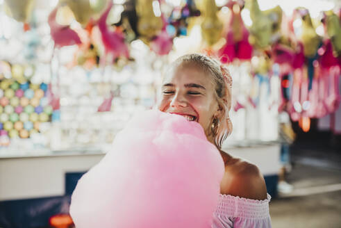 Nahaufnahme einer lächelnden jungen Frau mit geschlossenen Augen, die Zuckerwatte in einem Vergnügungspark hält - MIMFF00068