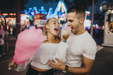 Cheerful young woman feeding cotton candy to boyfriend at amusement park - MIMFF00070