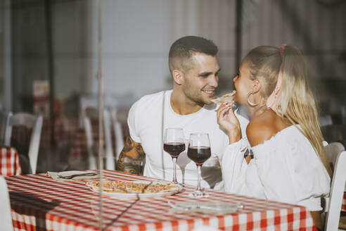 Lächelnder Mann, der seine Freundin beim Pizzaessen im Restaurant betrachtet, gesehen durch ein Fenster - MIMFF00071