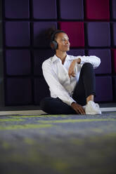 Porträt einer entspannten Geschäftsfrau, die im Büro auf dem Boden sitzt und mit drahtlosen Kopfhörern Musik hört - RBF07720