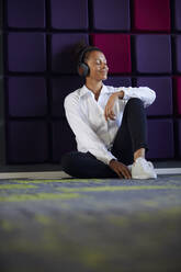 Porträt einer Geschäftsfrau mit geschlossenen Augen, die im Büro auf dem Boden sitzt und mit Kopfhörern Musik hört - RBF07721