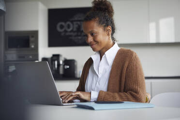 Lächelnde Geschäftsfrau, die am Tisch in der Küche sitzt und einen Laptop benutzt - RBF07753