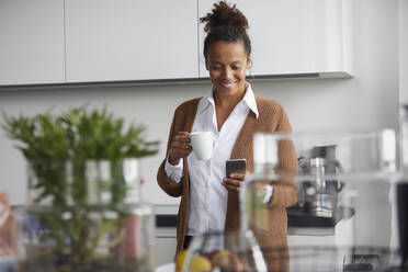 Portrait of smiling businesswoman standing in kitchen with cup of coffee looking at mobile phone - RBF07758