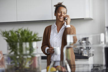 Portrait of smiling businesswoman on the phone standing in kitchen with cup of coffee looking at distance - RBF07759