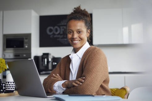 Porträt einer lächelnden Geschäftsfrau mit Laptop, die am Tisch in der Küche sitzt - RBF07762