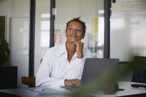 Porträt einer lächelnden Geschäftsfrau, die am Schreibtisch im Büro sitzt und in die Ferne schaut - RBF07771