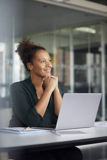 Porträt einer lächelnden Geschäftsfrau, die mit einem Laptop am Schreibtisch sitzt und in die Ferne schaut - RBF07775