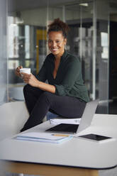 Porträt einer lächelnden Geschäftsfrau, die mit einer Tasse Kaffee am Schreibtisch sitzt und eine Pause macht - RBF07780