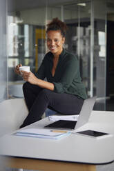 Portrait of smiling businesswoman sitting on desk with cup of coffee having a break - RBF07780