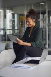 Smiling businesswoman sitting on desk using digital tablet - RBF07781