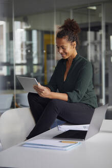 Lächelnde Geschäftsfrau, die auf einem Schreibtisch sitzt und ein digitales Tablet benutzt - RBF07781