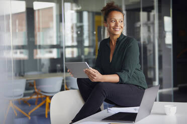 Porträt einer lächelnden Geschäftsfrau mit digitalem Tablet, die auf einem Schreibtisch sitzt und in die Ferne schaut - RBF07782
