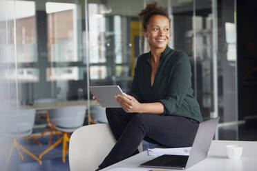 Portrait of smiling businesswoman with digital tablet sitting on desk looking at distance - RBF07782