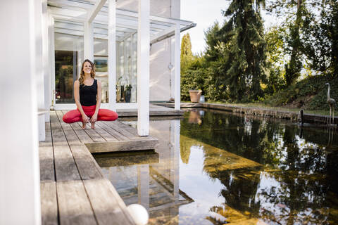 Mid adult woman meditating while crouching on hardwood floor by pond stock photo