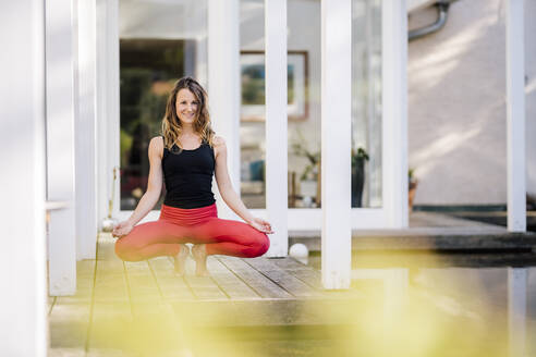 Smiling woman meditating while crouching on hardwood floor against house - DAWF01629