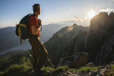 Männlicher Wanderer mit Rucksack mit Blick auf die Berge bei Sonnenuntergang, Orobie, Lecco, Italien - MCVF00451