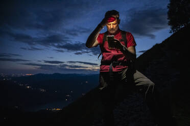 Male hiker checking map over smart phone while standing on mountain at night, Orobie, Lecco, Italy - MCVF00459