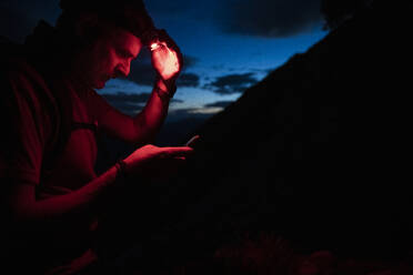 Man with headlamp checking map over smart phone at night, Orobie, Lecco, Italy - MCVF00462