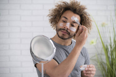 Close-up of young man applying facial mask while looking in mirror at home - SNF00336