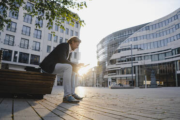 Businessman sitting on a bench in the city at sunset using tablet - JOSEF00815