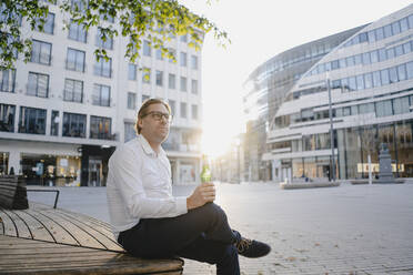 Geschäftsmann sitzt auf einer Bank in der Stadt bei Sonnenuntergang mit einer Flasche Bier - JOSEF00816