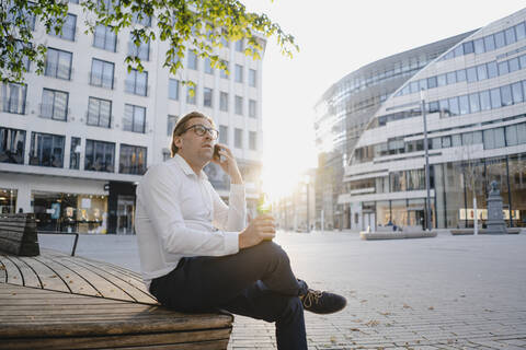 Geschäftsmann sitzt auf einer Bank in der Stadt bei Sonnenuntergang und telefoniert, lizenzfreies Stockfoto