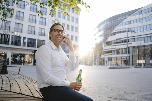 Businessman sitting on a bench in the city at sunset talking on the phone - JOSEF00829