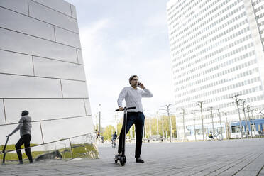 Geschäftsmann mit Kick-Scooter und Smartphone spiegelt sich in Gebäudefassade - JOSEF00875