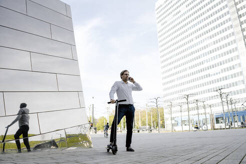 Geschäftsmann mit Kick-Scooter und Smartphone spiegelt sich in Gebäudefassade - JOSEF00875