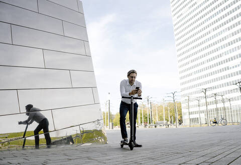 Geschäftsmann mit Kick-Scooter und Smartphone spiegelt sich in Gebäudefassade, lizenzfreies Stockfoto