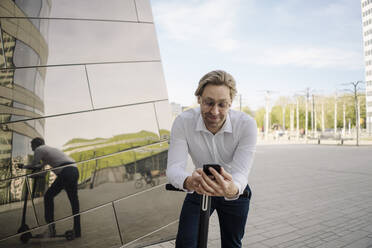 Businessman with kick scooter and smartphone reflected in building facade - JOSEF00877