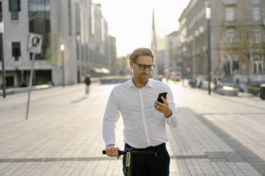 Businessman with kick scooter using smartphone in the city - JOSEF00896