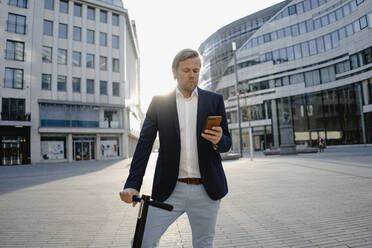 Businessman with kick scooter using smartphone in the city at sunset - JOSEF00912