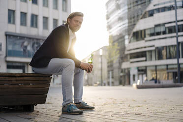 Geschäftsmann sitzt auf einer Bank in der Stadt bei Sonnenuntergang mit einer Flasche Bier - JOSEF00917