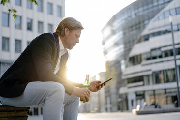 Geschäftsmann sitzt auf einer Bank in der Stadt bei Sonnenuntergang mit einer Flasche Bier und Smartphone - JOSEF00918