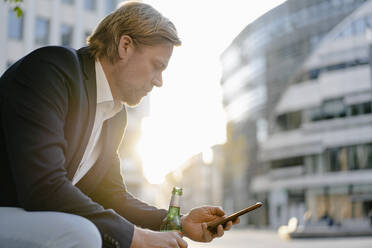 Geschäftsmann sitzt auf einer Bank in der Stadt bei Sonnenuntergang mit einer Flasche Bier und Smartphone - JOSEF00919